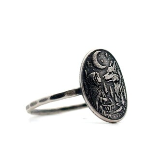 May include: Silver ring featuring an oval pendant with a detailed engraving. The design depicts a hooded figure facing a wolf under a crescent moon and stars, with a forest in the background. The ring band has a hammered texture.