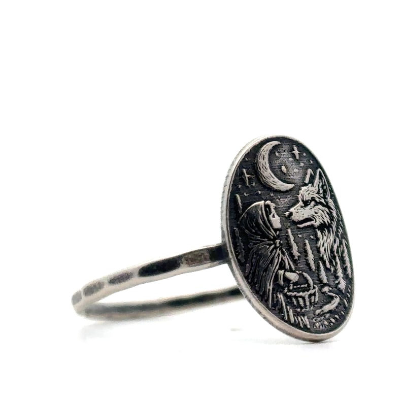 May include: Silver ring featuring an oval pendant with a detailed engraving. The design depicts a hooded figure facing a wolf under a crescent moon and stars, with a forest in the background. The ring band has a hammered texture.