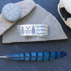 May include: A silver bracelet with an engraved design of trees, a deer, and branches. The bracelet is on a gray stone surface.
