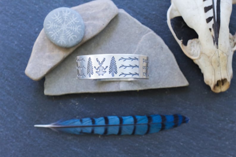 May include: A silver bracelet with an engraved design of trees, a deer, and branches. The bracelet is on a gray stone surface.