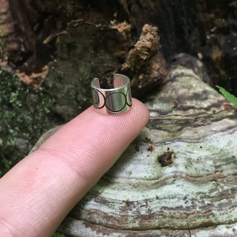 May include: Silver ear cuff with a design of multiple moon phases. The cuff is on a finger.