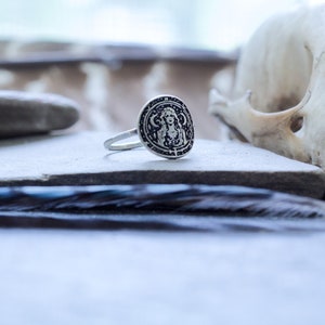 May include: A silver ring with a circular design featuring a woman's face and intricate details. The ring is on a gray surface with a feather in the background.