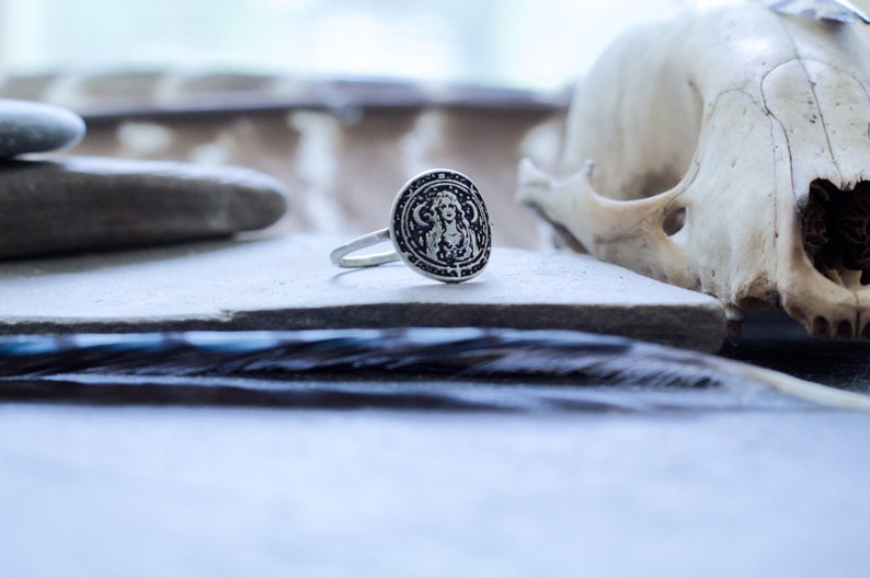 May include: A silver ring with a circular design featuring a woman's face and intricate details. The ring is on a gray surface with a feather in the background.