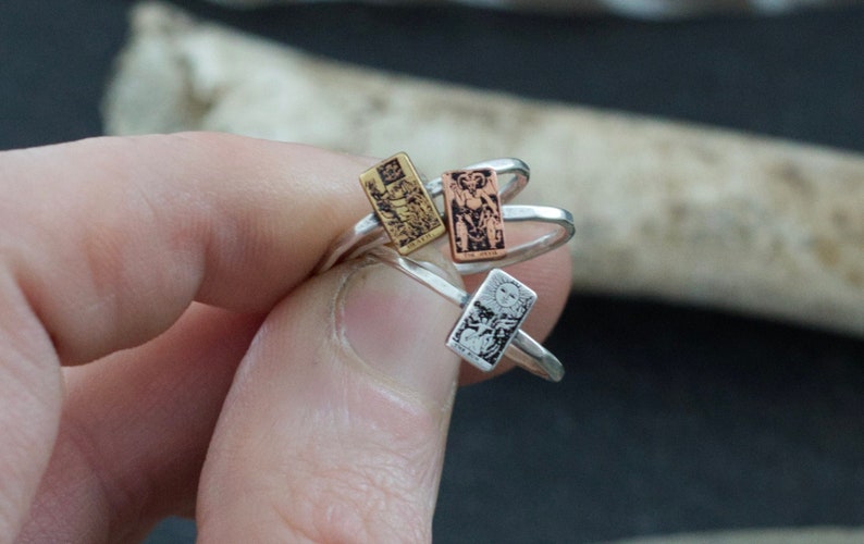 May include: Three silver rings with different tarot card designs. The rings are stacked on top of each other. The top ring features a gold card, the middle ring features a copper card, and the bottom ring features a silver card.