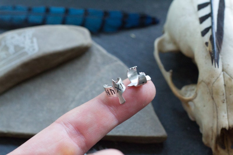 May include: Two silver rings in the shape of a bird with the word "XIX" inscribed on one ring. The rings are on a finger.