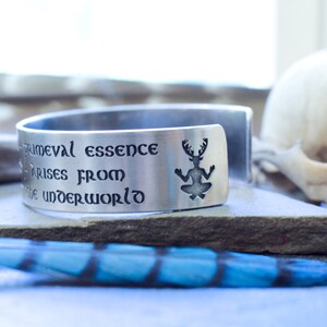 May include: A silver cuff bracelet with an engraved quote: "Primeval essence arises from the underworld." The quote is surrounded by a silhouette of a creature with antlers.