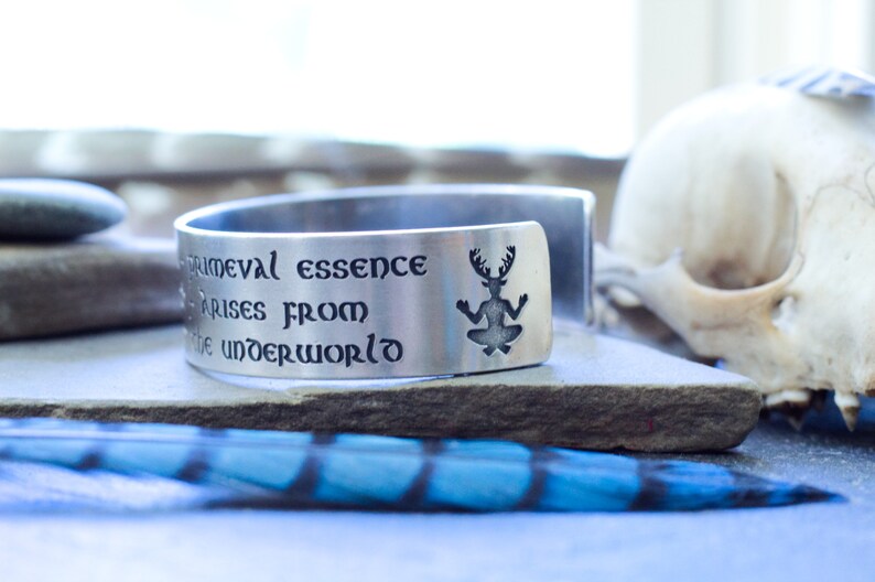 May include: A silver cuff bracelet with an engraved quote: "Primeval essence arises from the underworld." The quote is surrounded by a silhouette of a creature with antlers.