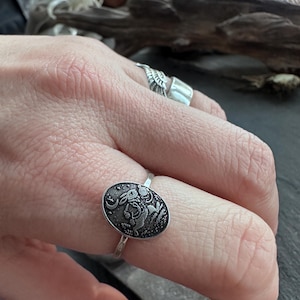 May include: A silver ring featuring an oval-shaped face with a detailed engraving of a rabbit, moon, and stars. The ring is worn on a finger, showcasing the intricate design and craftsmanship. The ring's band is thin and simple, complementing the detailed face.