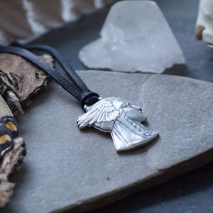May include: A silver pendant necklace featuring a profile of a woman wearing a headdress with feathers. The necklace is on a black leather cord and is resting on a gray stone surface.