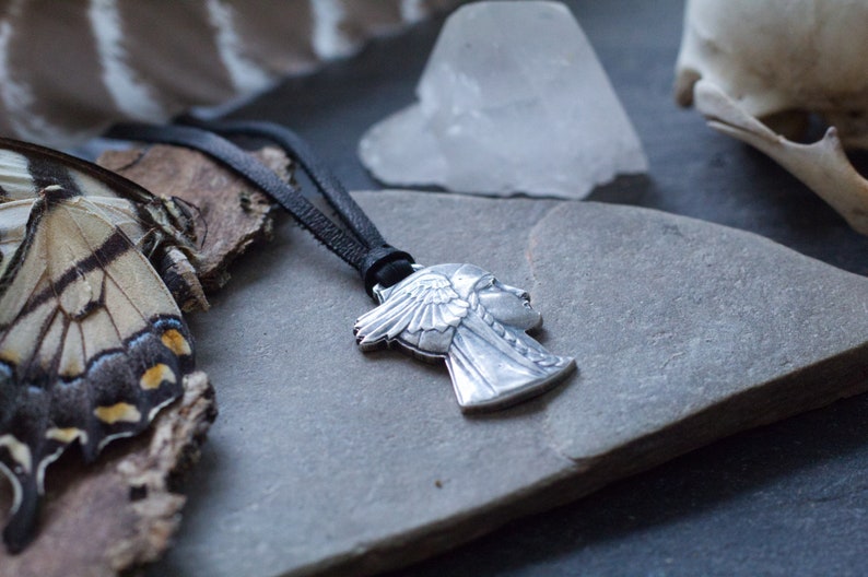 May include: A silver pendant necklace featuring a profile of a woman wearing a headdress with feathers. The necklace is on a black leather cord and is resting on a gray stone surface.