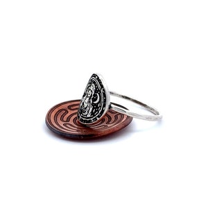 May include: A silver ring with a detailed design of a woman's face and a crescent moon. The ring is sitting on a copper-colored dish with a maze pattern.
