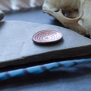May include: A copper-colored metal disc with a circular design etched into the surface. The disc is resting on a gray stone surface.