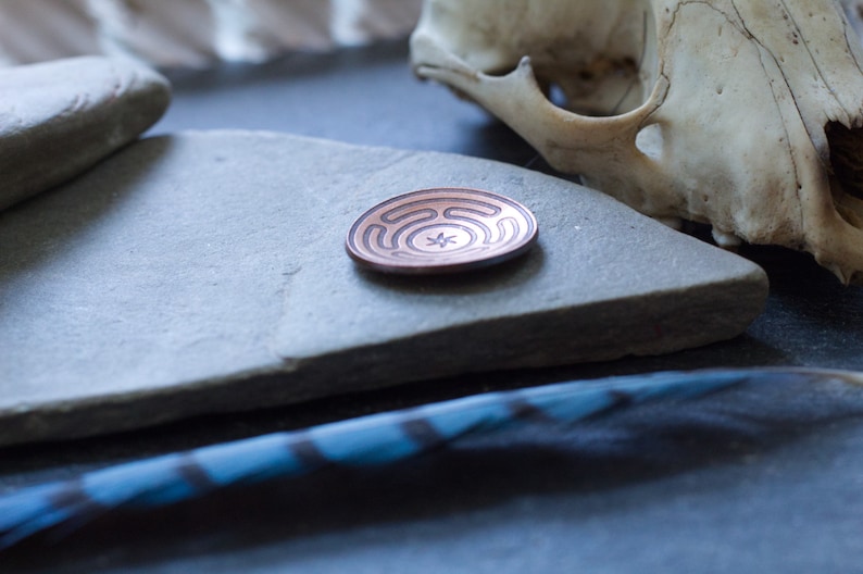 May include: A copper-colored metal disc with a circular design etched into the surface. The disc is resting on a gray stone surface.