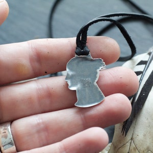May include: Silver pendant necklace with a profile of a woman's face. The pendant is on a black leather cord. The pendant has the text "Made in Italy" on the back.