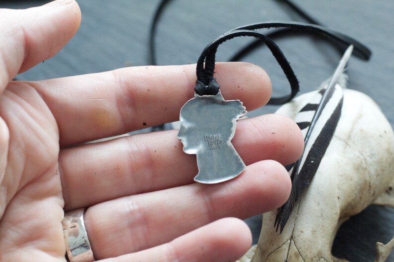 May include: Silver pendant necklace with a profile of a woman's face. The pendant is on a black leather cord. The pendant has the text "Made in Italy" on the back.