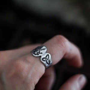 May include: A silver ring with a Celtic knot design. The ring is worn on a person's finger.