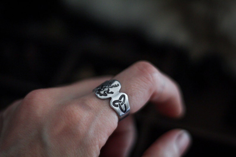 May include: A silver ring with a Celtic knot design. The ring is worn on a person's finger.