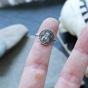May include: A silver ring with a circular design featuring a woman's face surrounded by a circle of symbols.