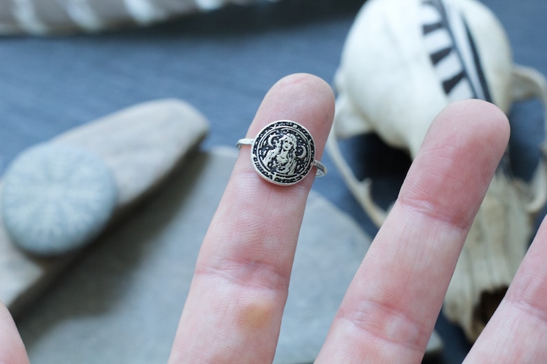May include: A silver ring with a circular design featuring a woman's face surrounded by a circle of symbols.