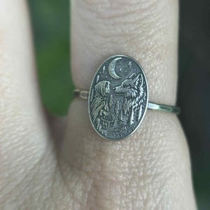 May include: Silver ring with an oval pendant featuring a detailed engraving of a wolf, a figure, a crescent moon, and stars. The ring is worn on a finger, showcasing the intricate design and craftsmanship of the jewelry.