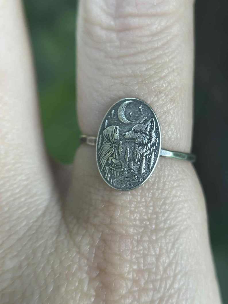 May include: Silver ring with an oval pendant featuring a detailed engraving of a wolf, a figure, a crescent moon, and stars. The ring is worn on a finger, showcasing the intricate design and craftsmanship of the jewelry.