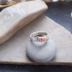 May include: A silver ring with three stacked bands, each featuring a different rune symbol. The runes are engraved in black on silver, copper, and gold bands. The ring is displayed on a gray stone.