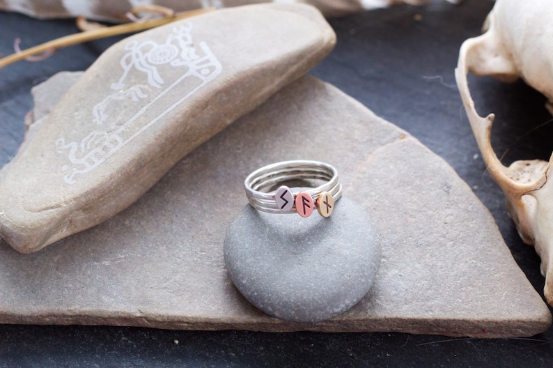 May include: A silver ring with three stacked bands, each featuring a different rune symbol. The runes are engraved in black on silver, copper, and gold bands. The ring is displayed on a gray stone.