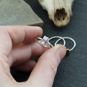 May include: A silver ring with a textured rectangular design. The ring is on a finger.