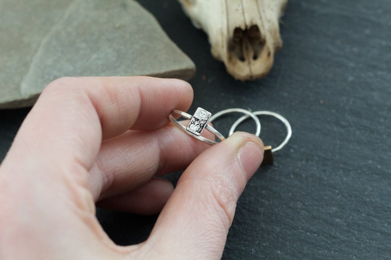 May include: A silver ring with a textured rectangular design. The ring is on a finger.