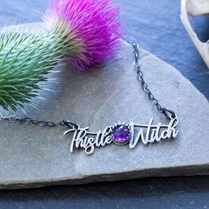 May include: A silver necklace with a purple gemstone and the words "Thistle Witch" written in cursive.