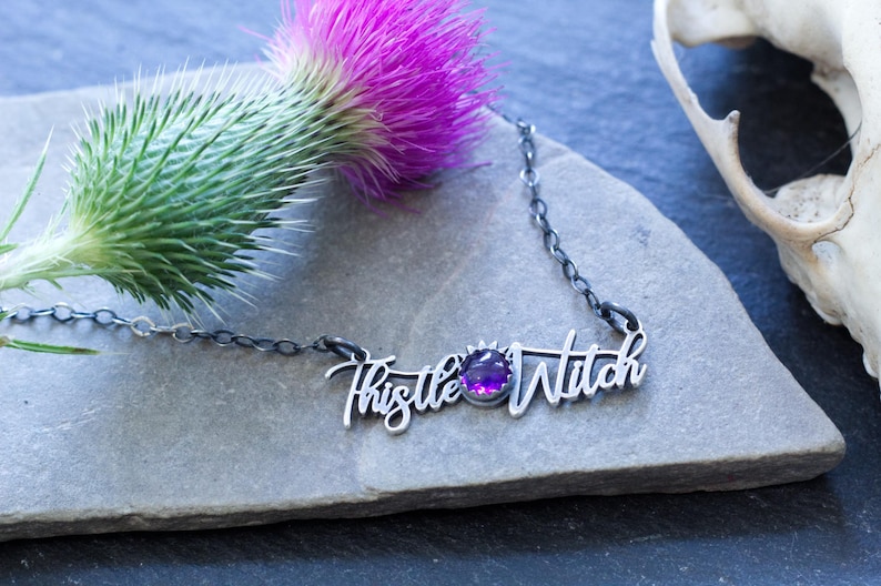 May include: A silver necklace with a purple gemstone and the words "Thistle Witch" written in cursive.