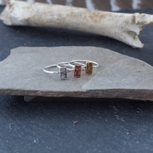 May include: Four silver rings with a rectangular design on a gray stone surface. The rings are stacked on top of each other.