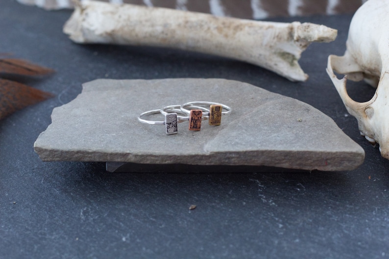 May include: Four silver rings with a rectangular design on a gray stone surface. The rings are stacked on top of each other.