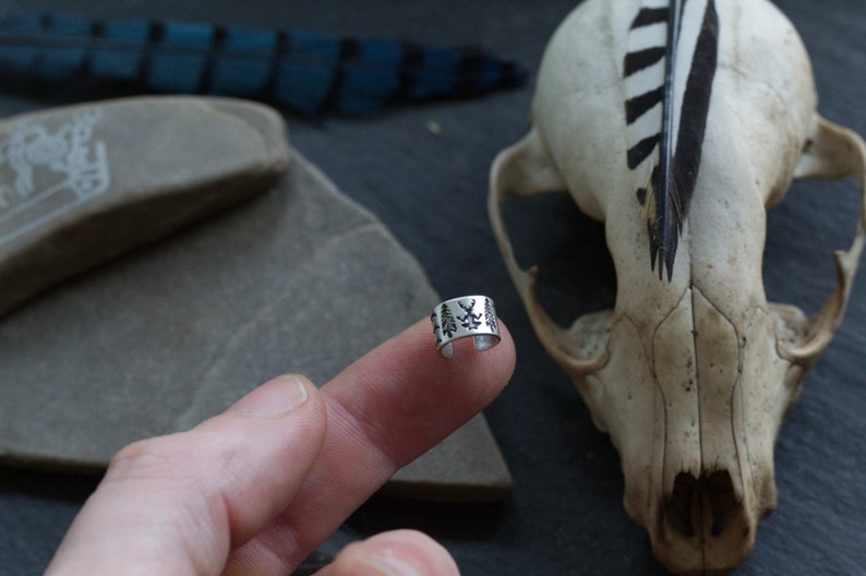 May include: A silver ear cuff with a design of small trees. The cuff is on a finger, and a skull with a feather is in the background.