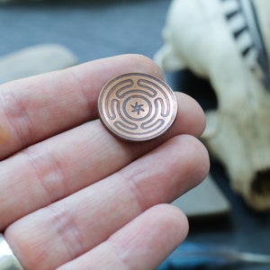 May include: A copper-colored metal disc with a circular design etched into the surface. The design features a central star with concentric circles radiating outwards.