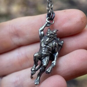 May include: A silver pendant necklace featuring a detailed depiction of a horned, humanoid figure with a beard and a basket on its back. The pendant is suspended from a delicate chain, showcasing a gothic or mythological theme.
