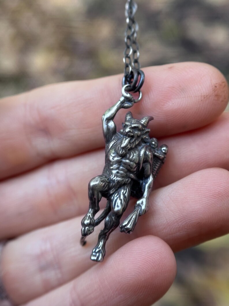 May include: A silver pendant necklace featuring a detailed depiction of a horned, humanoid figure with a beard and a basket on its back. The pendant is suspended from a delicate chain, showcasing a gothic or mythological theme.