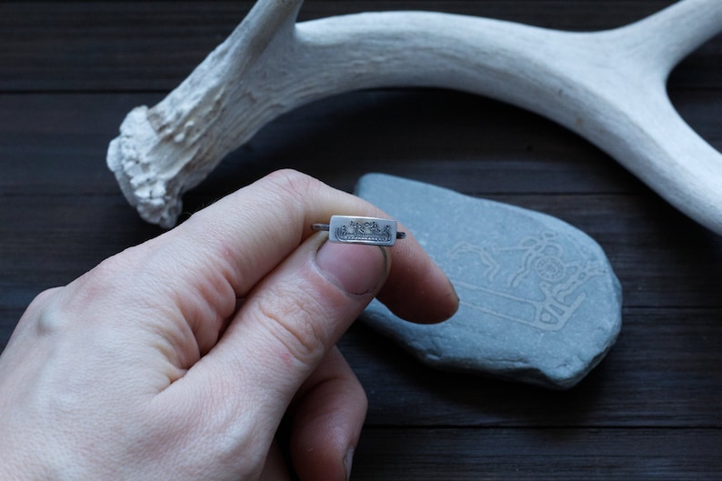 May include: A silver ring with a rectangular face featuring a detailed landscape design. The ring is held in a hand, showcasing the intricate craftsmanship. The background includes a white antler and a gray stone.