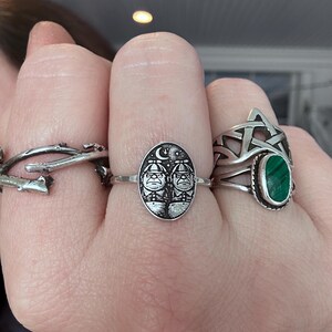 May include: A hand displays three silver rings. One ring features a twig design, another has an oval face with two figures under a crescent moon and stars, and the third has a pentagram with a green oval stone.