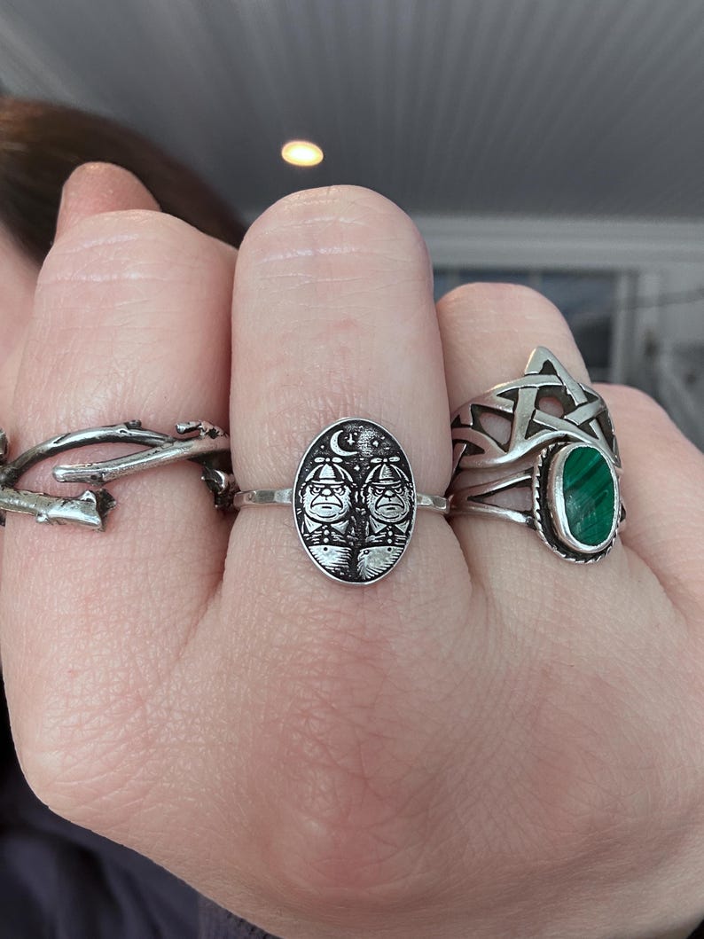 May include: A hand displays three silver rings. One ring features a twig design, another has an oval face with two figures under a crescent moon and stars, and the third has a pentagram with a green oval stone.
