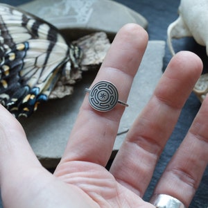 May include: A silver ring featuring a circular design with a maze-like pattern and a central star. The ring is worn on a finger, with a butterfly and skull in the background. The ring's design is detailed and intricate.