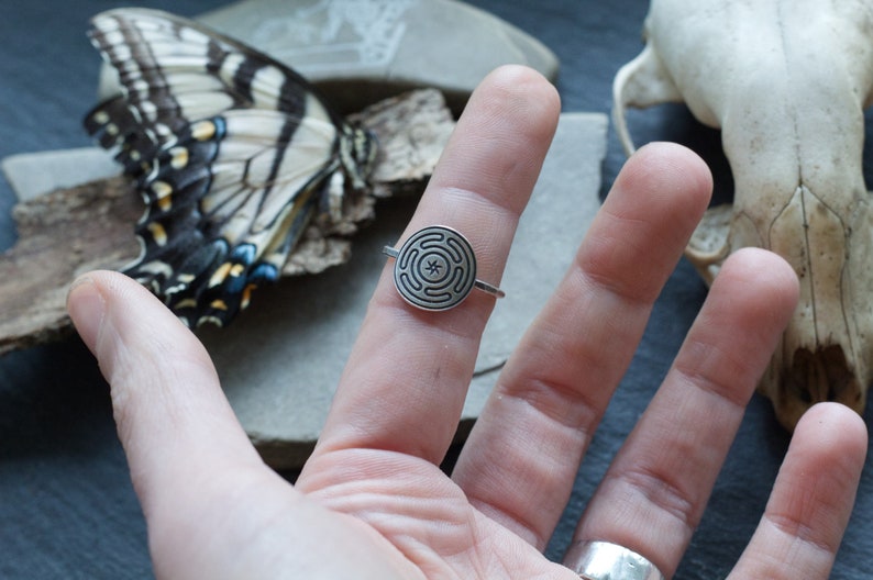 May include: A silver ring featuring a circular design with a maze-like pattern and a central star. The ring is worn on a finger, with a butterfly and skull in the background. The ring's design is detailed and intricate.