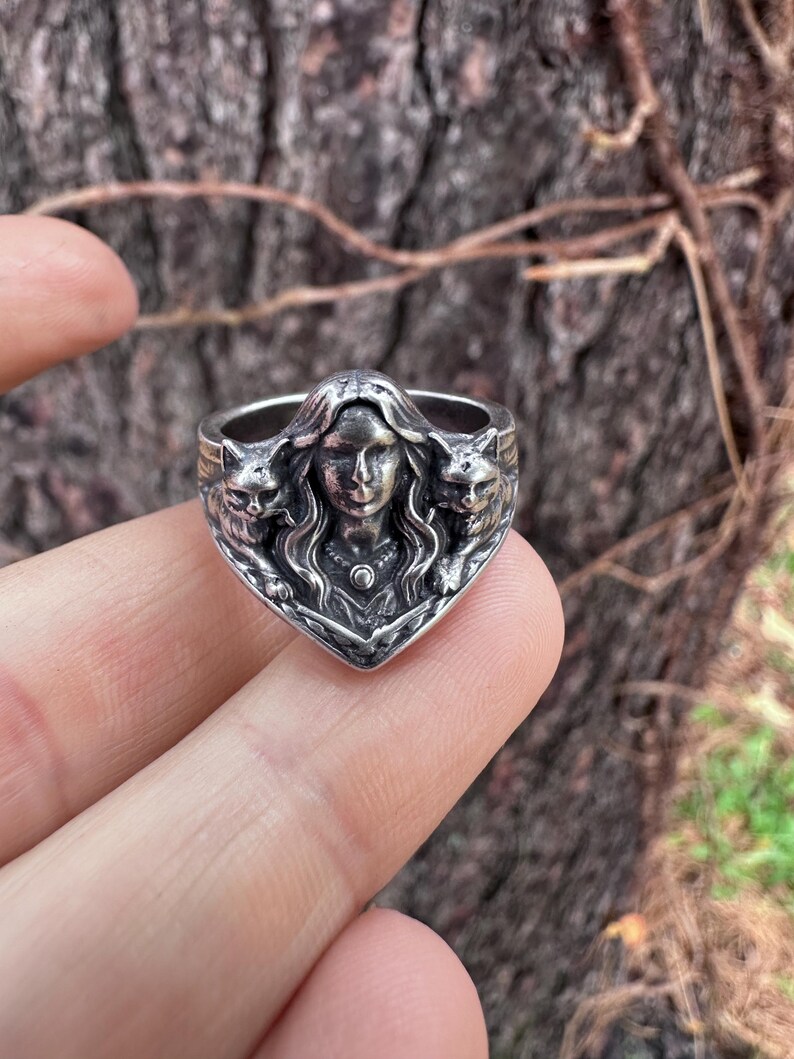 K&ouml;nnte beinhalten: Ein silberner Ring mit einer zentralen weiblichen Figur, flankiert von zwei Katzenfiguren. Der Ring hat aufwendige Details und einen Vintage-Look, ideal f&uuml;r Liebhaber von einzigartigem Schmuck.