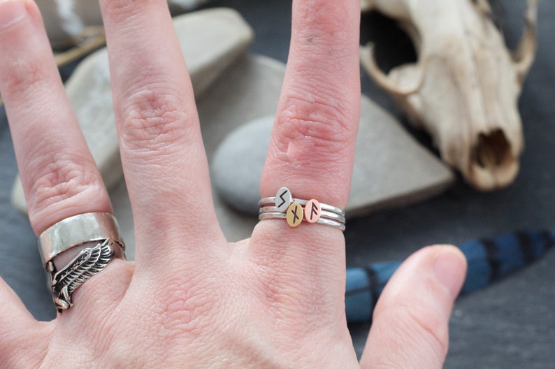 May include: A hand wearing three silver stacking rings with small round charms. The charms have different runes engraved on them. The runes are "S", "X", and "F".