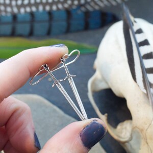 Peut inclure: Une paire de boucles d'oreilles en forme d'&eacute;p&eacute;e argent&eacute;es avec un design simple. Les boucles d'oreilles sont suspendues &agrave; l'oreille d'une personne.