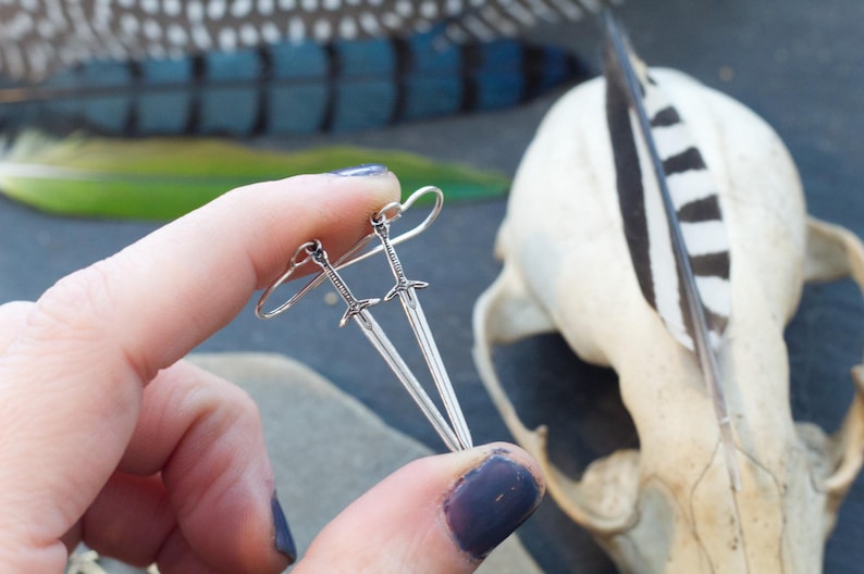 Peut inclure: Une paire de boucles d'oreilles en forme d'&eacute;p&eacute;e argent&eacute;es avec un design simple. Les boucles d'oreilles sont suspendues &agrave; l'oreille d'une personne.