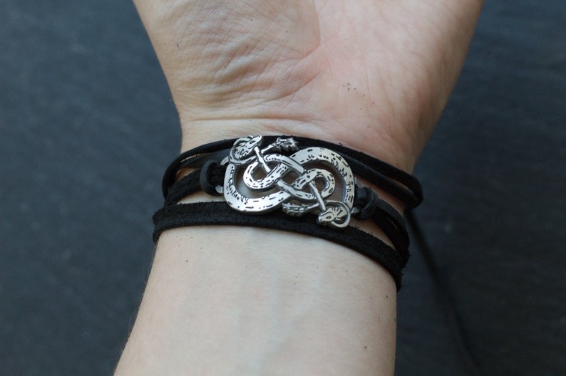 May include: A black leather bracelet with a silver snake knot charm. The bracelet is adjustable and has a braided leather design.