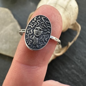 May include: A silver ring featuring an oval medallion with a detailed Medusa design. The ring's band is simple, and the Medusa image includes a crescent moon and stars. The ring is shown on a finger against a dark background.
