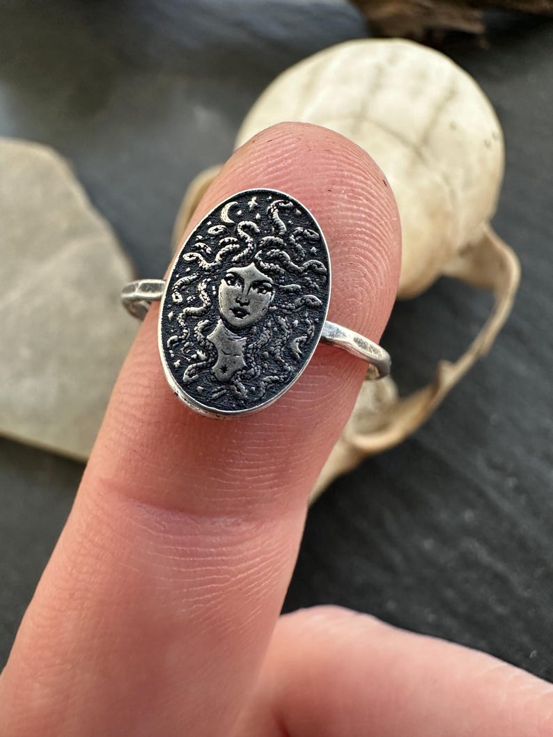 May include: A silver ring featuring an oval medallion with a detailed Medusa design. The ring's band is simple, and the Medusa image includes a crescent moon and stars. The ring is shown on a finger against a dark background.