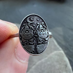 May include: Silver ring with an oval face featuring two stylized figures wearing hats, under a crescent moon and stars. The figures have stern expressions. The ring is held against a neutral background.